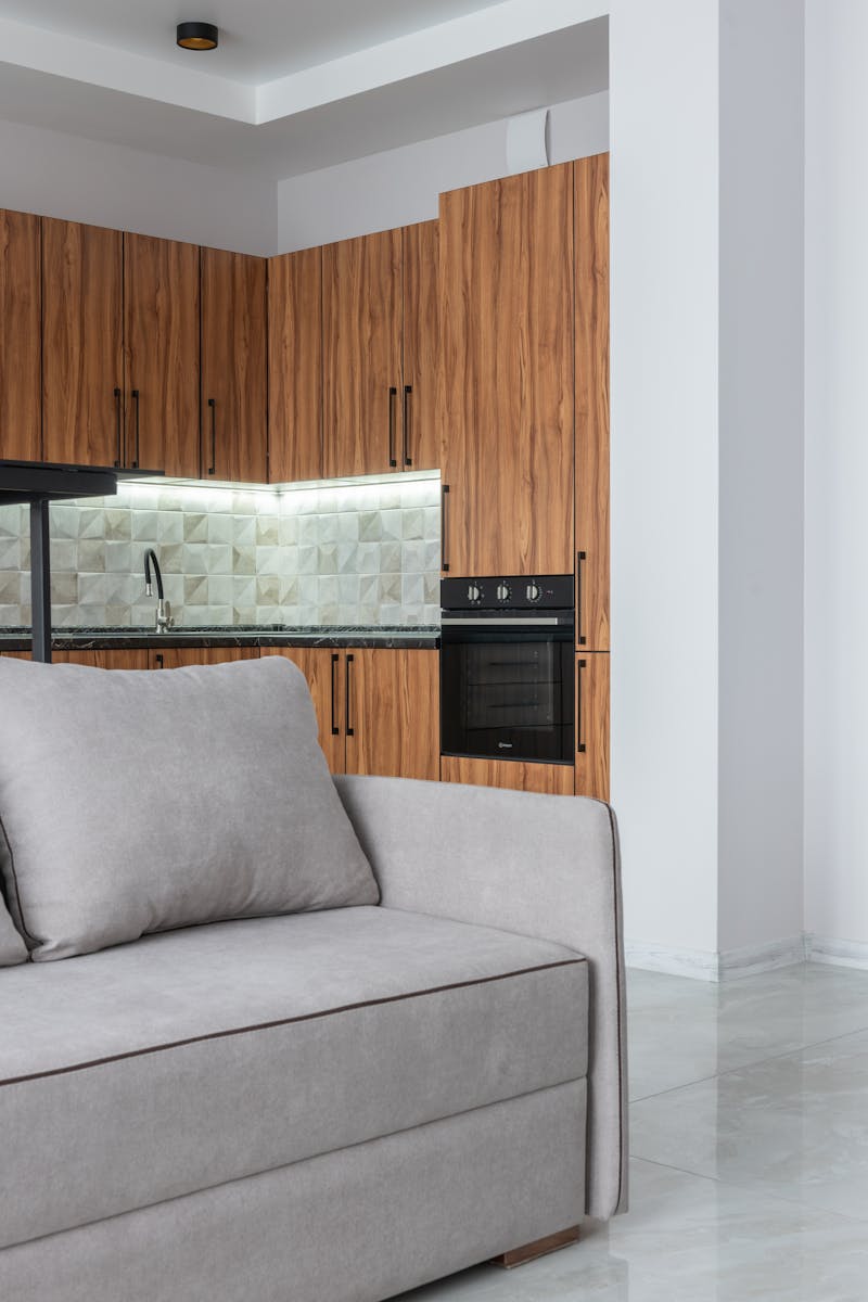 Interior of contemporary spacious light room with kitchen counter with modern oven near wooden cabinets and comfortable sofa at home